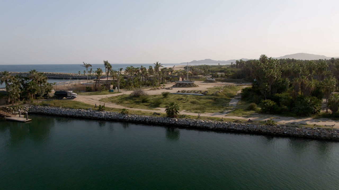 Aerial view over dark green water toward a rocky shoreline with palms and distant ocean.