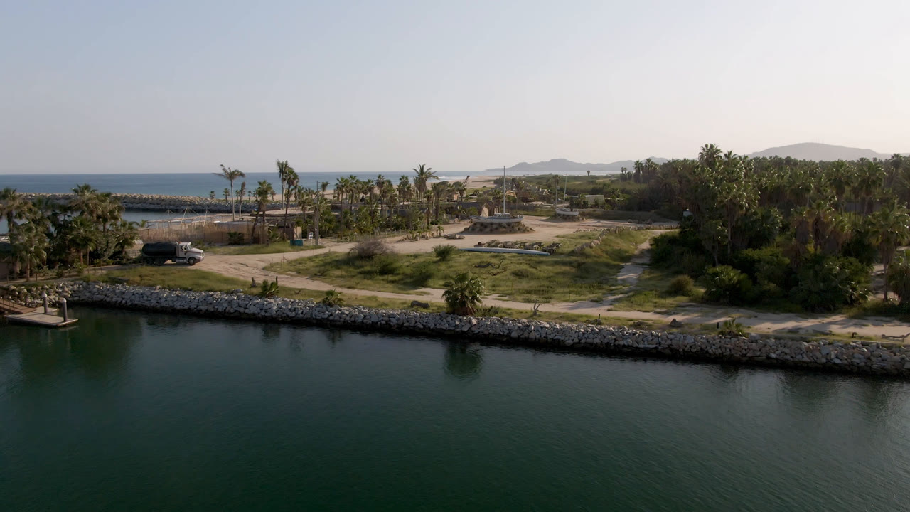 A drone frame in Puerto Los Cabos shows sheltered green water below, a rocky bank across the middle, sandy access tracks, palm trees, distant ocean, low hills, and no visible people.