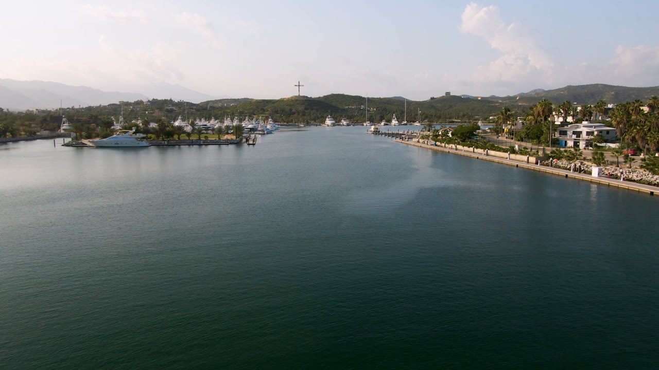 Wide aerial frame over Puerto Los Cabos water with a yacht near the left shore, a palm-lined waterfront on the right, hazy hills behind, and no visible people.