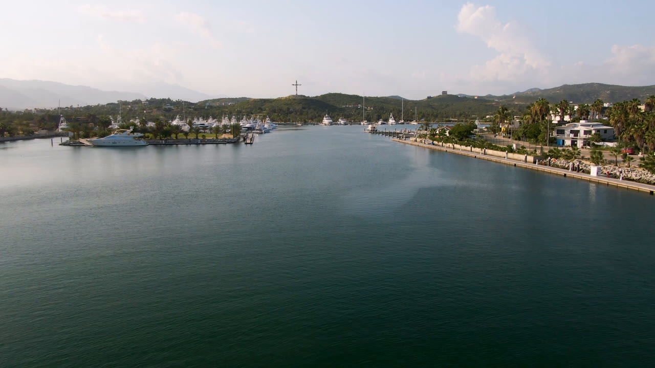 High wide Puerto Los Cabos frame with calm water in front, a large yacht near the left marina edge, palm trees and buildings to the right, distant mountains, and no visible people.