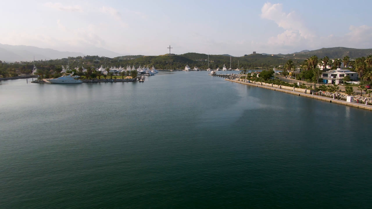 Aerial frame from Puerto Los Cabos showing open green-blue water in the foreground, yachts and docks along the shore, palm trees, low buildings, distant hills, scattered clouds, and no visible people.