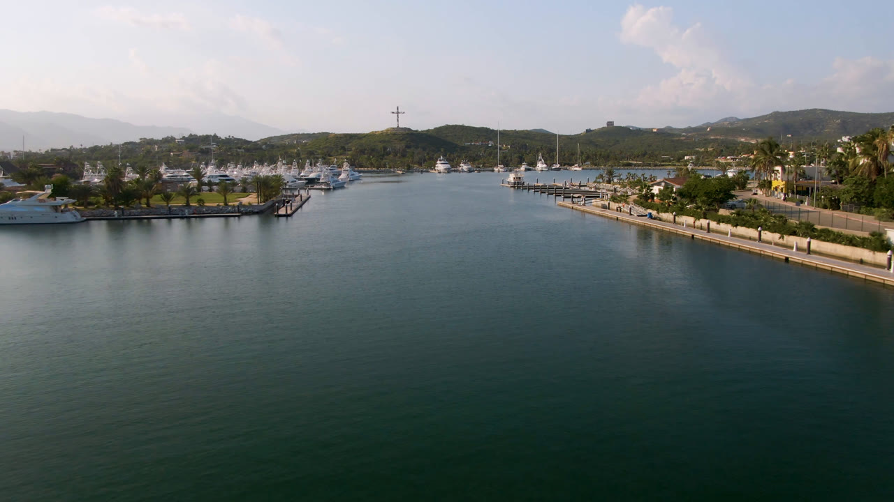 Aerial frame over Puerto Los Cabos marina water, with docks and boats along both sides, low hills in the distance, soft clouds above, and no visible people.