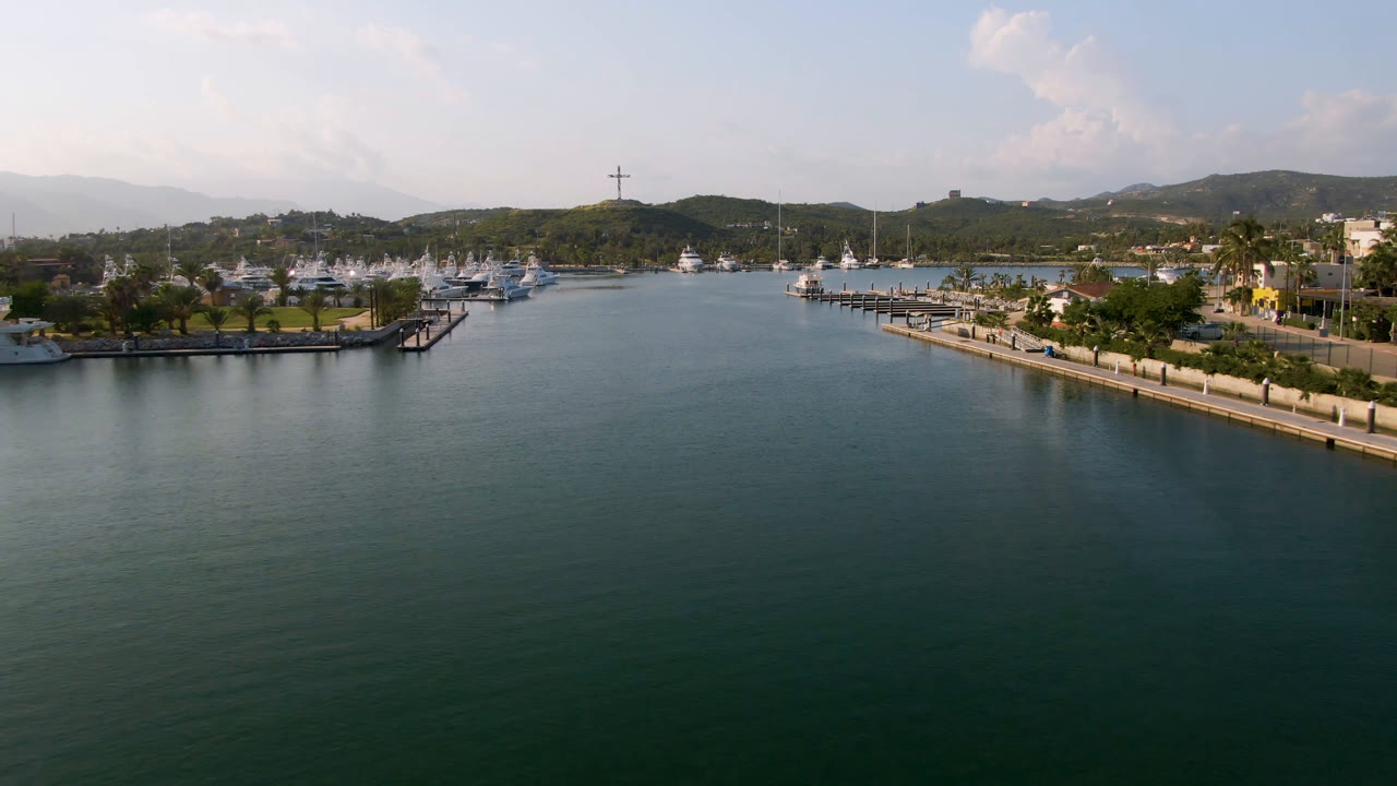 Aerial view of Puerto Los Cabos water with a long dock and moored boats on the right, more boats on the left shore, distant hills, and no visible people.