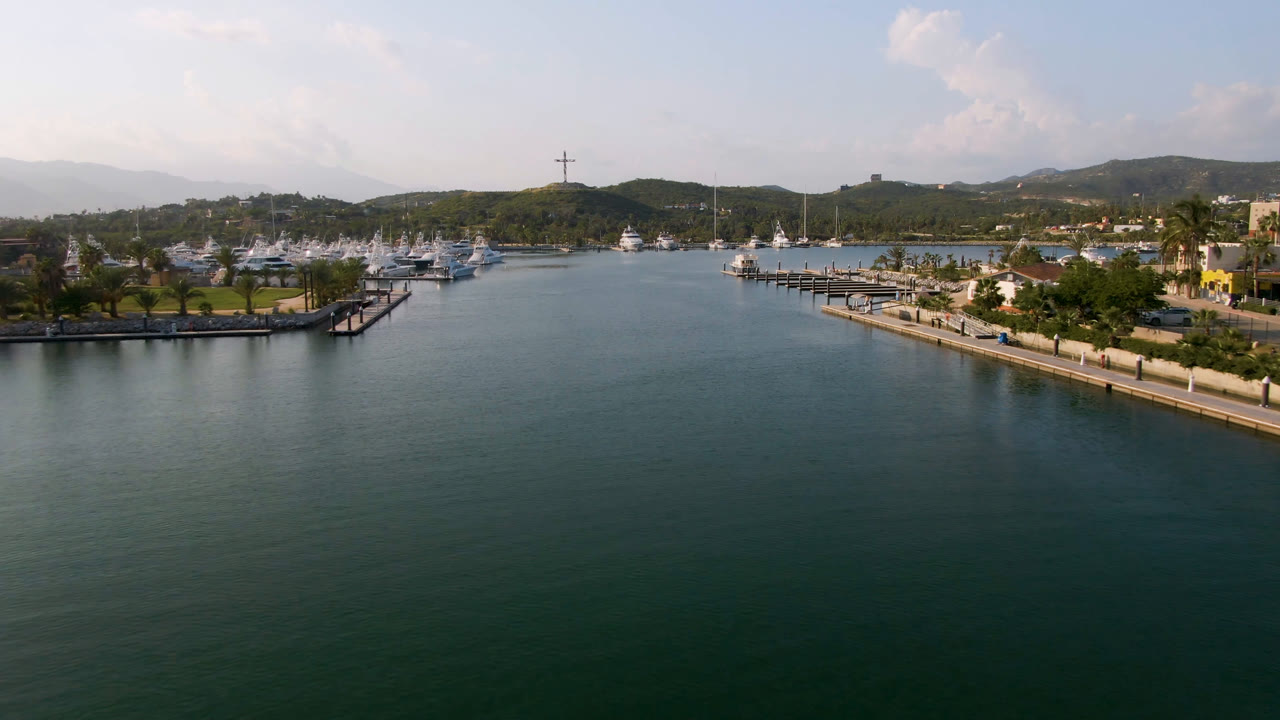 Marina water with boats, docks, palm trees, shoreline buildings, and hills under bright sky.