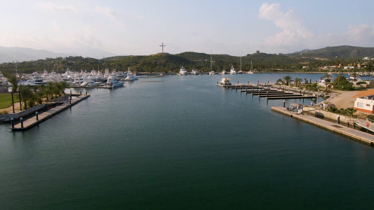 Aerial view of marina water, docks, boats, palms, hills, and a cross on a hill.