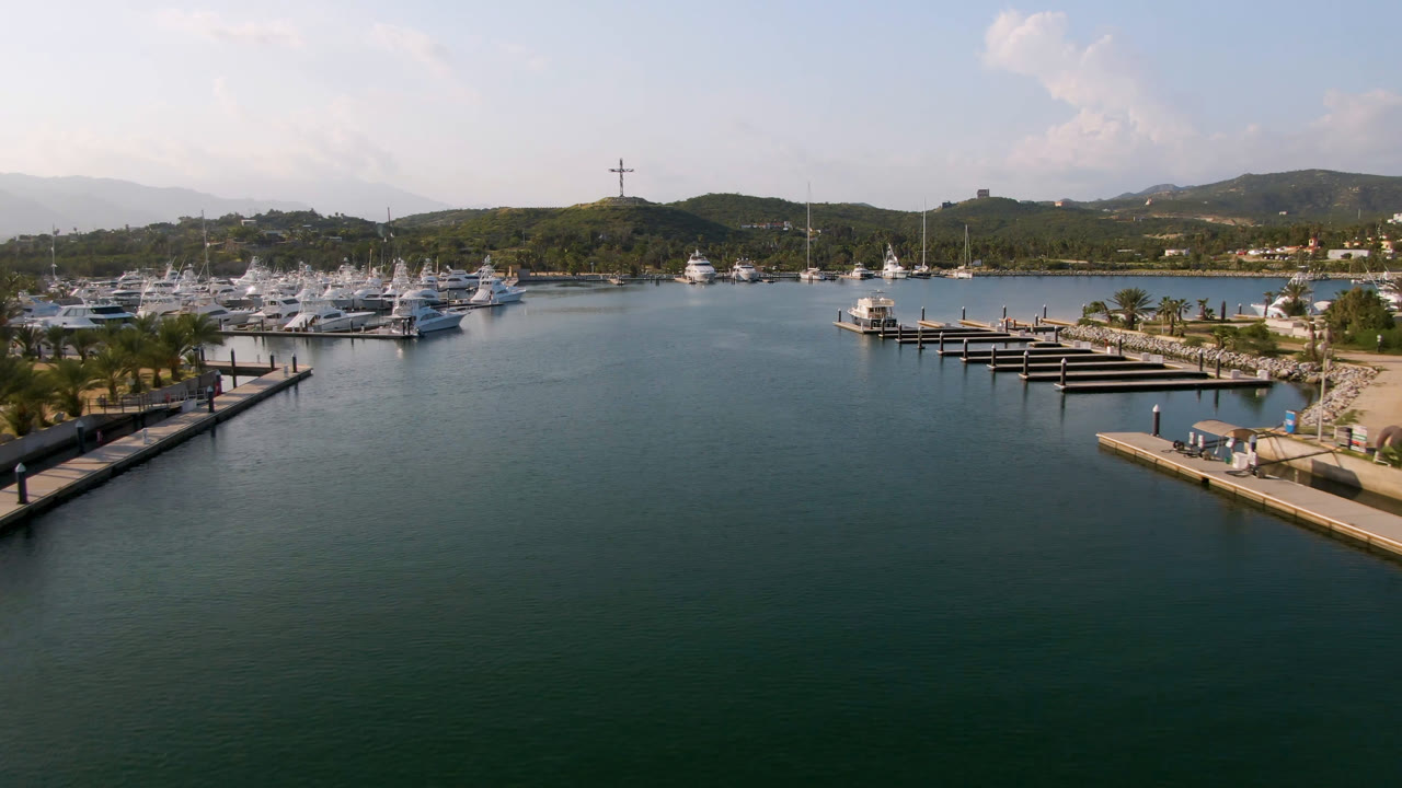 Aerial view in Puerto Los Cabos showing broad marina water, a central dock system with empty slips, yachts clustered along the far shore, palms on the waterfront, distant hills, a visible cross on a ridge, and no visible people.