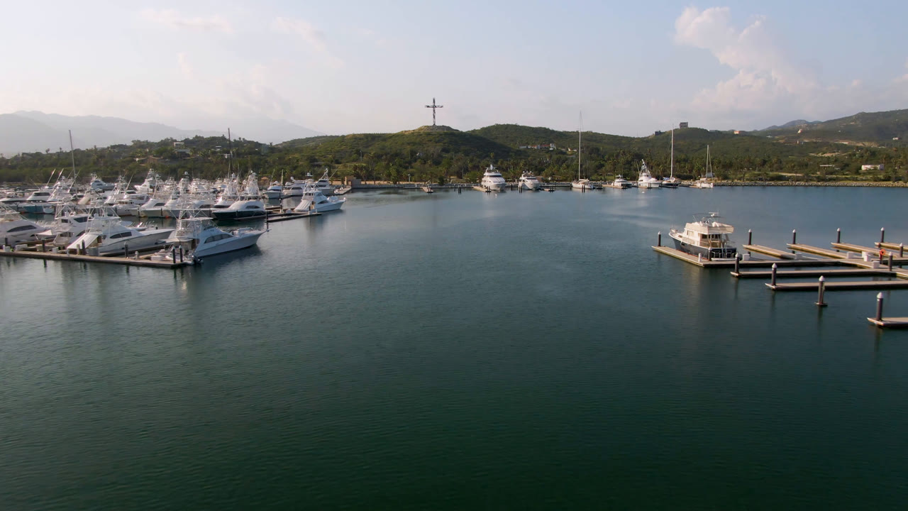 Aerial frame of Puerto Los Cabos marina with rows of white boats at left, open dark water in front, green hills and hazy mountains behind, and no visible people.