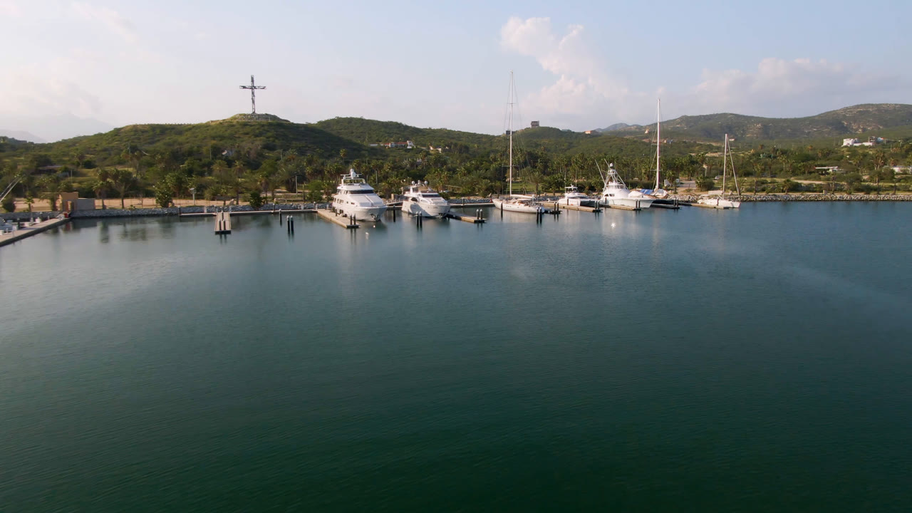 Wide aerial view of marina boats, calm water, and hills with a large cross.