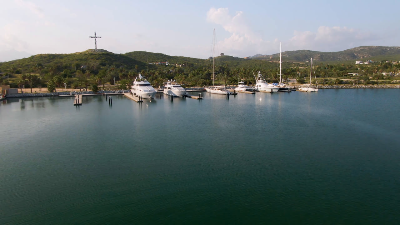 At 0:01, the Puerto Los Cabos aerial view holds on docked yachts and sailboats across the midground, with broad calm water in front, low hills behind, a hilltop cross, and no visible people.