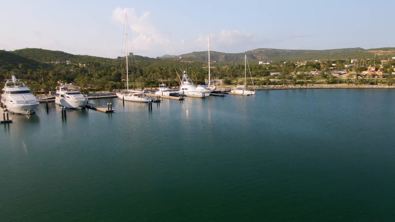 Puerto Los Cabos aerial frame showing docked motor yachts and sailboats slightly farther right in the composition, with calm water, hillside buildings, and no visible people.