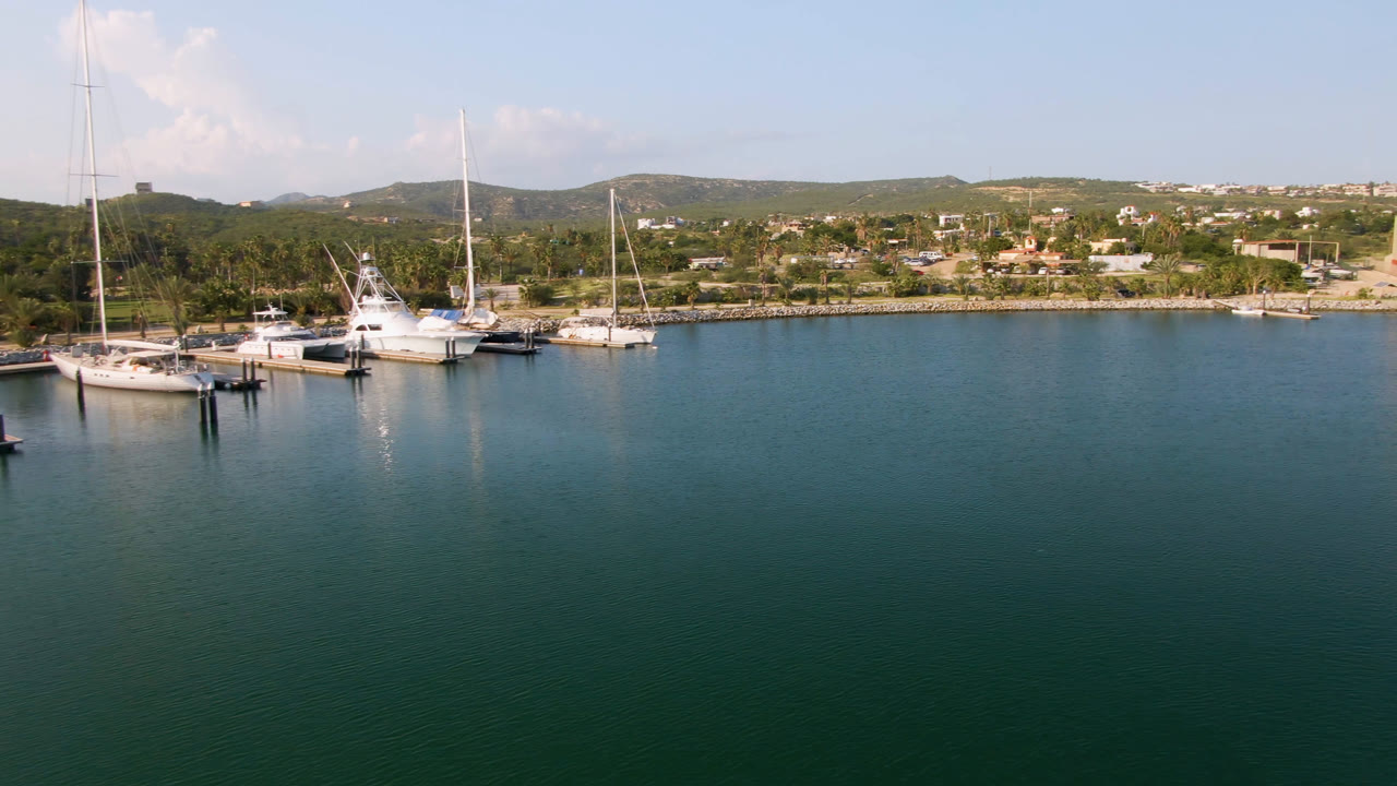 Opening frame of Puerto Los Cabos with green-blue harbor water in the foreground, several white boats docked at left, shoreline buildings and hills behind them, and no visible people.