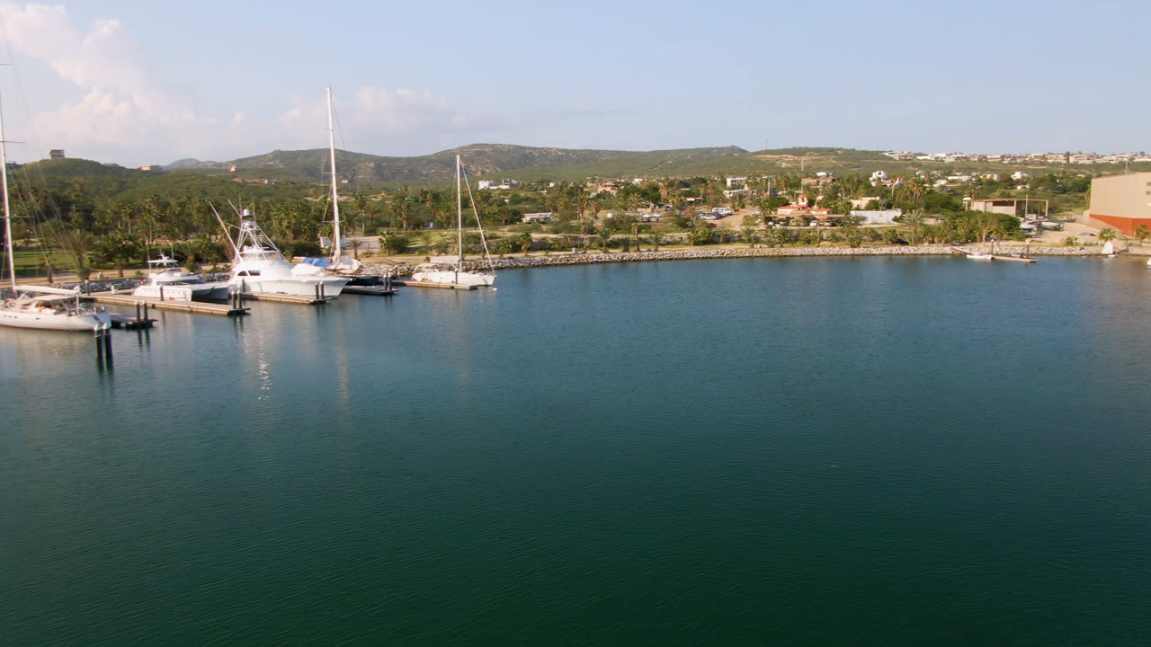 Puerto Los Cabos harbor frame with docked white yachts and sailboats left of center, smooth water across the foreground, shoreline development and hills in the distance, and no visible people.