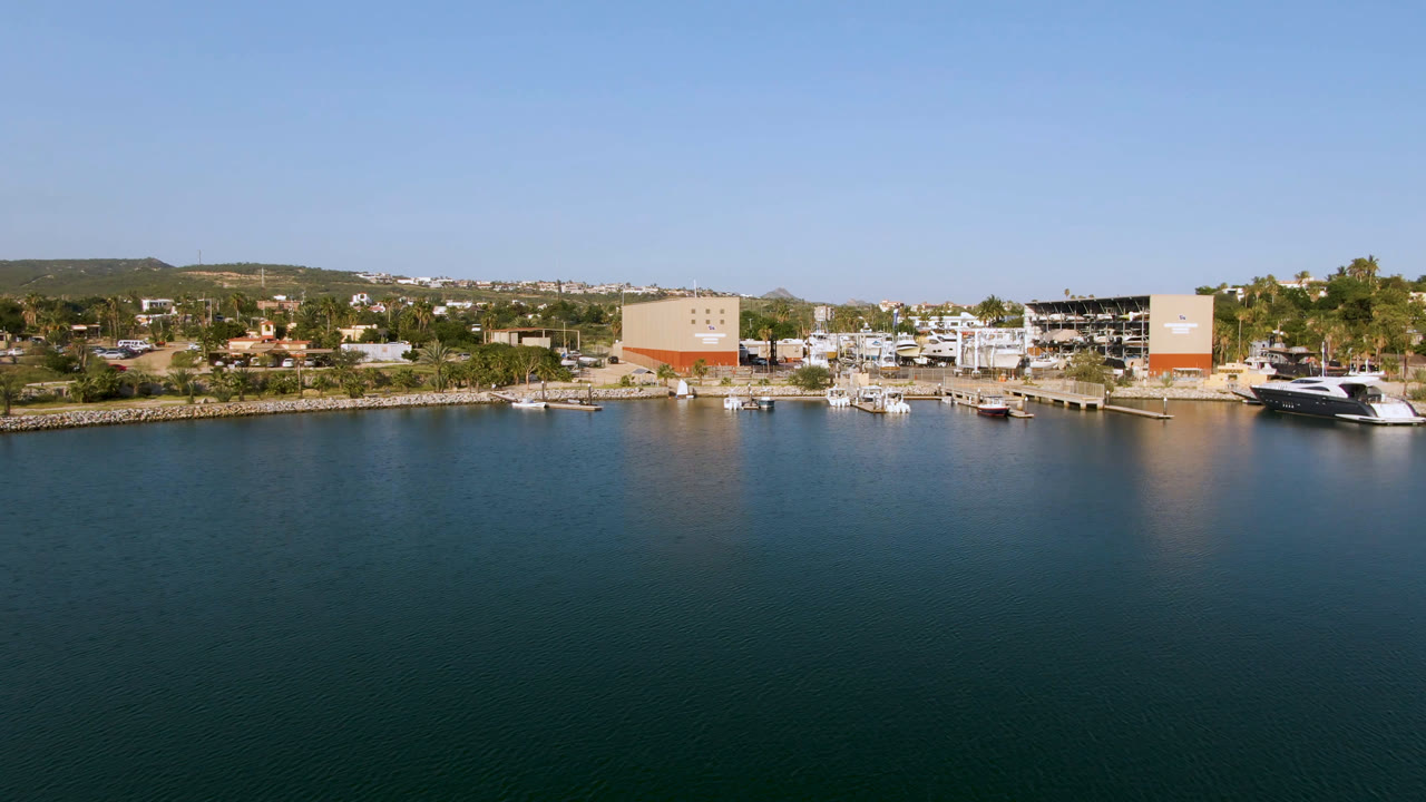 Aerial frame from Puerto Los Cabos showing dark blue-green water in the foreground, marina boats and shoreline buildings beyond, distant low hills, clear sky, and no visible people.