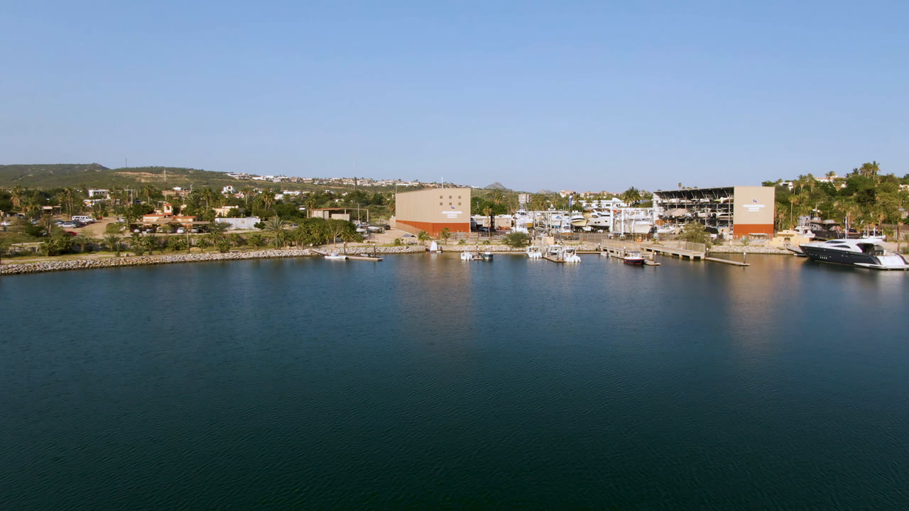 Puerto Los Cabos aerial view with open water across the lower frame, clustered marina boats near the shore, trees, tan and white buildings, distant hills, and no visible people.