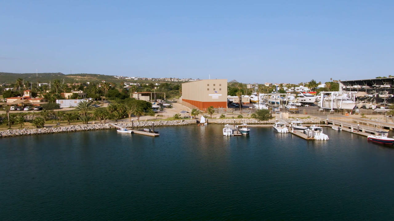 Elevated view of Puerto Los Cabos marina with dark water in the foreground, several docked boats, a palm-lined shore, a large beige and orange building, and no visible people.