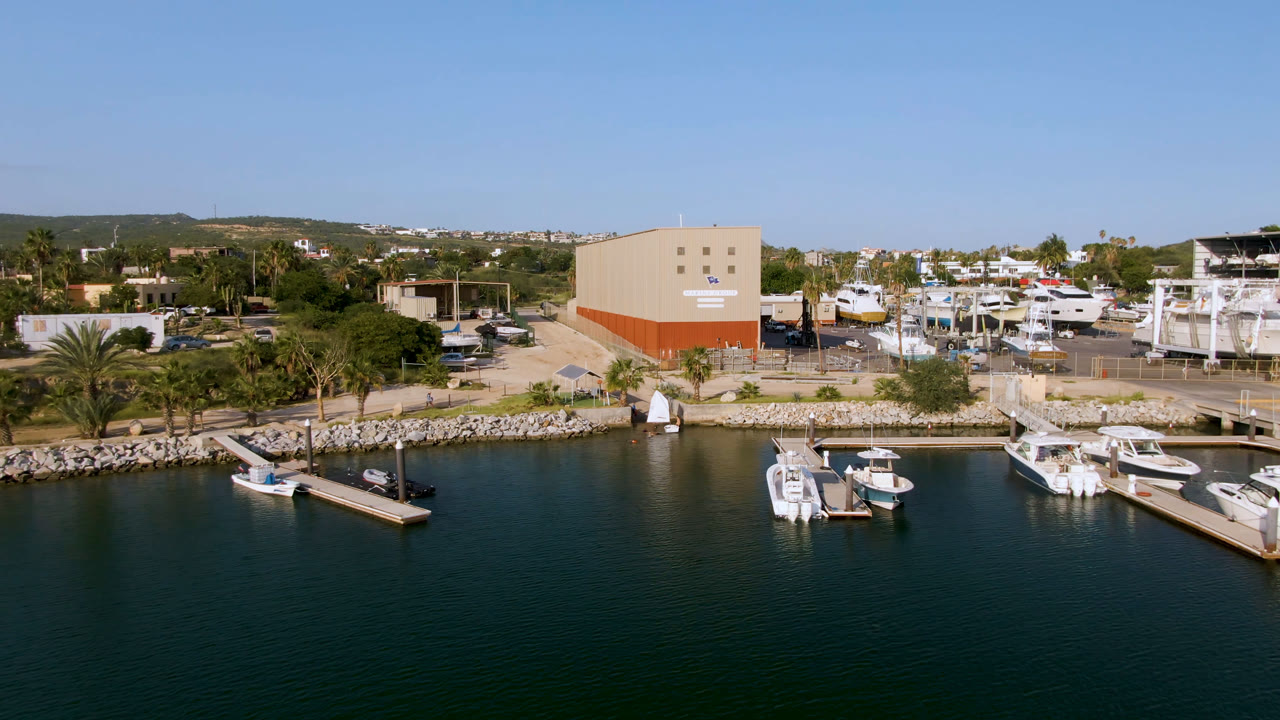 Aerial frame over Puerto Los Cabos marina showing dark green water, floating docks, several white boats, palms, beige buildings, and no visible people at the start of the clip.