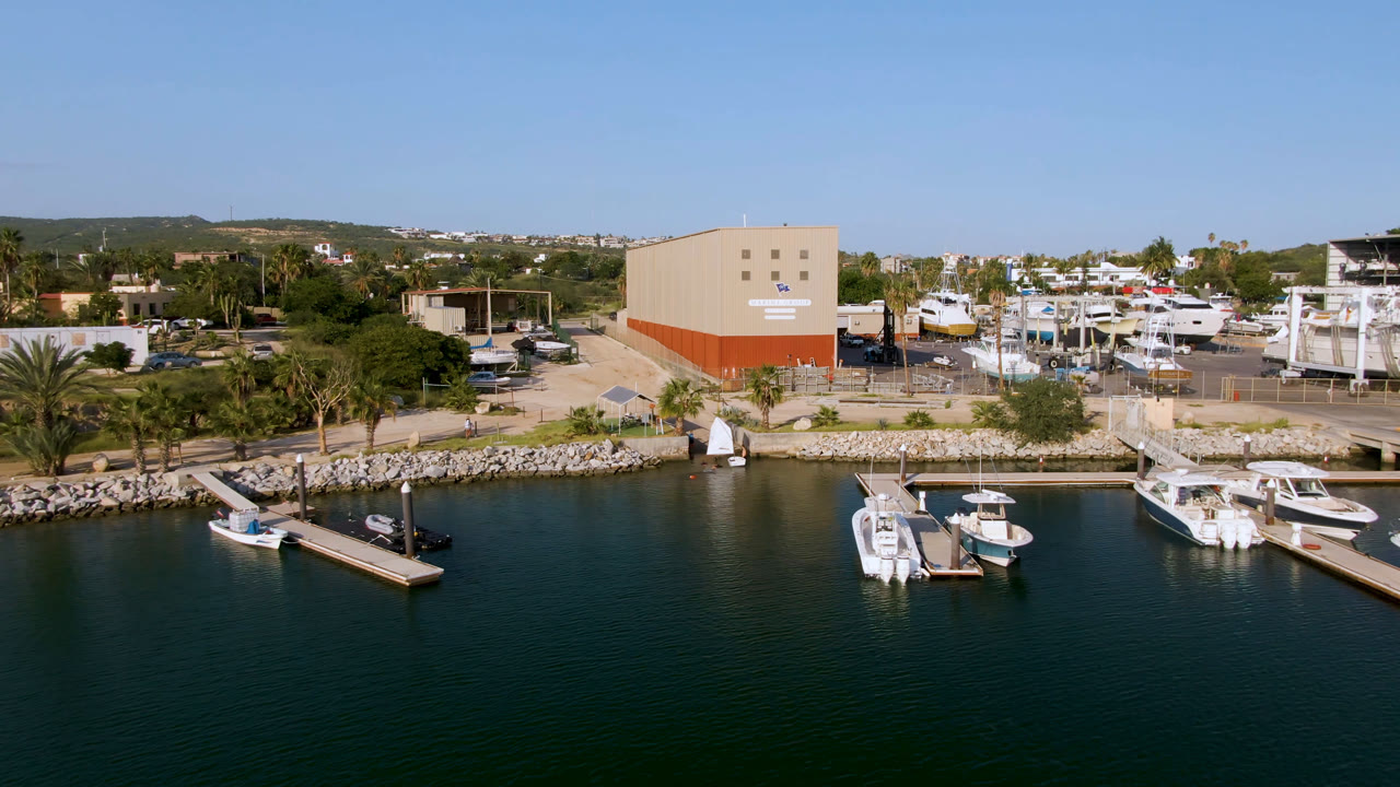 Puerto Los Cabos marina aerial with a pair of small boats near the foreground docks, shoreline palms, boatyard structures, hills beyond, and no visible people.