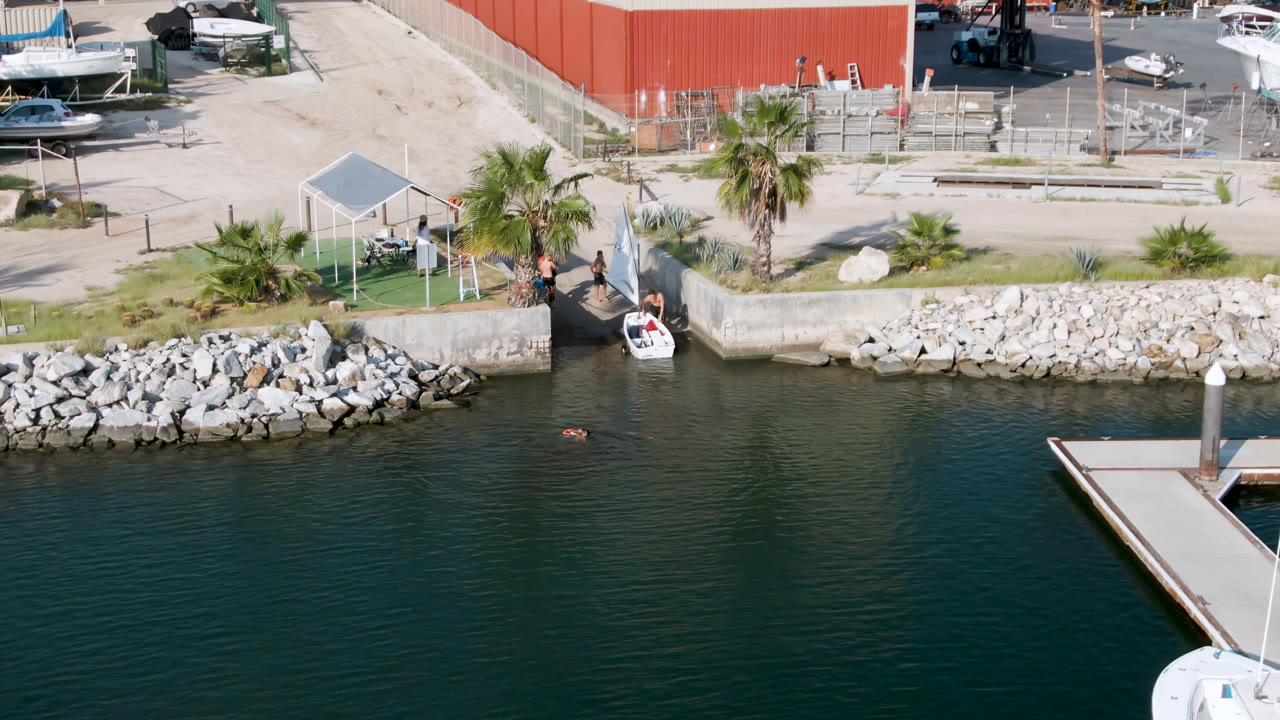 Overhead view in San Jose Del Cabo, Mexico showing dark marina water, a floating dock corner, a rock-lined shore, palms, and a small white boat at the ramp with no identifiable people.