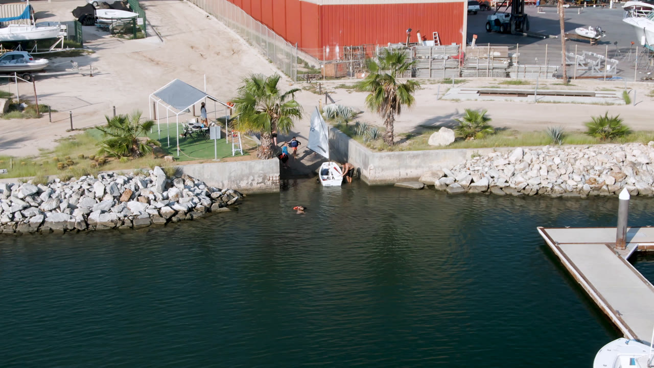 Overhead marina edge with dock, rocks, palm trees, boats, and red building