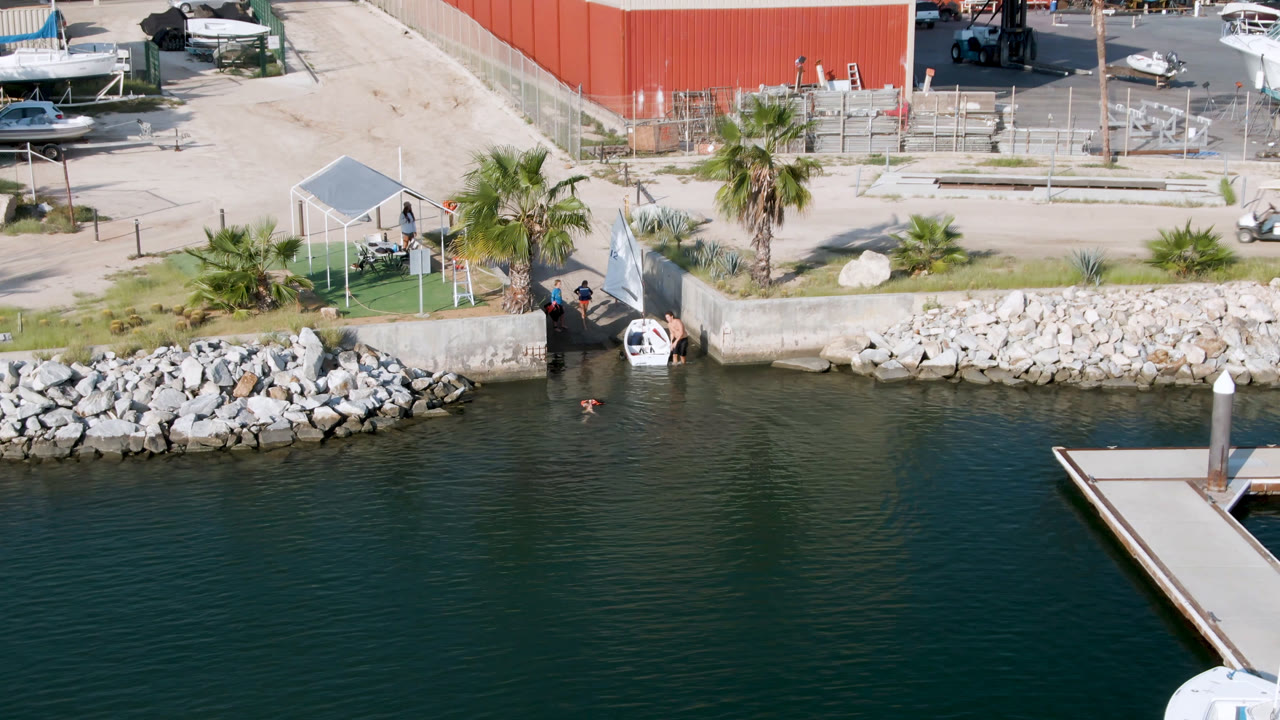 Aerial frame from San Jose Del Cabo, Mexico, showing the dock corner over dark water, a rock-lined shore, a small covered area, palm trees, parked boats, and small people near the water access.