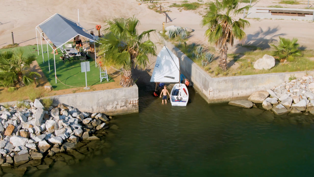 Aerial frame from San Jose Del Cabo, Mexico, with the white boat held near the seawall, people clustered close to the water, and a palm-lined picnic spot behind it.