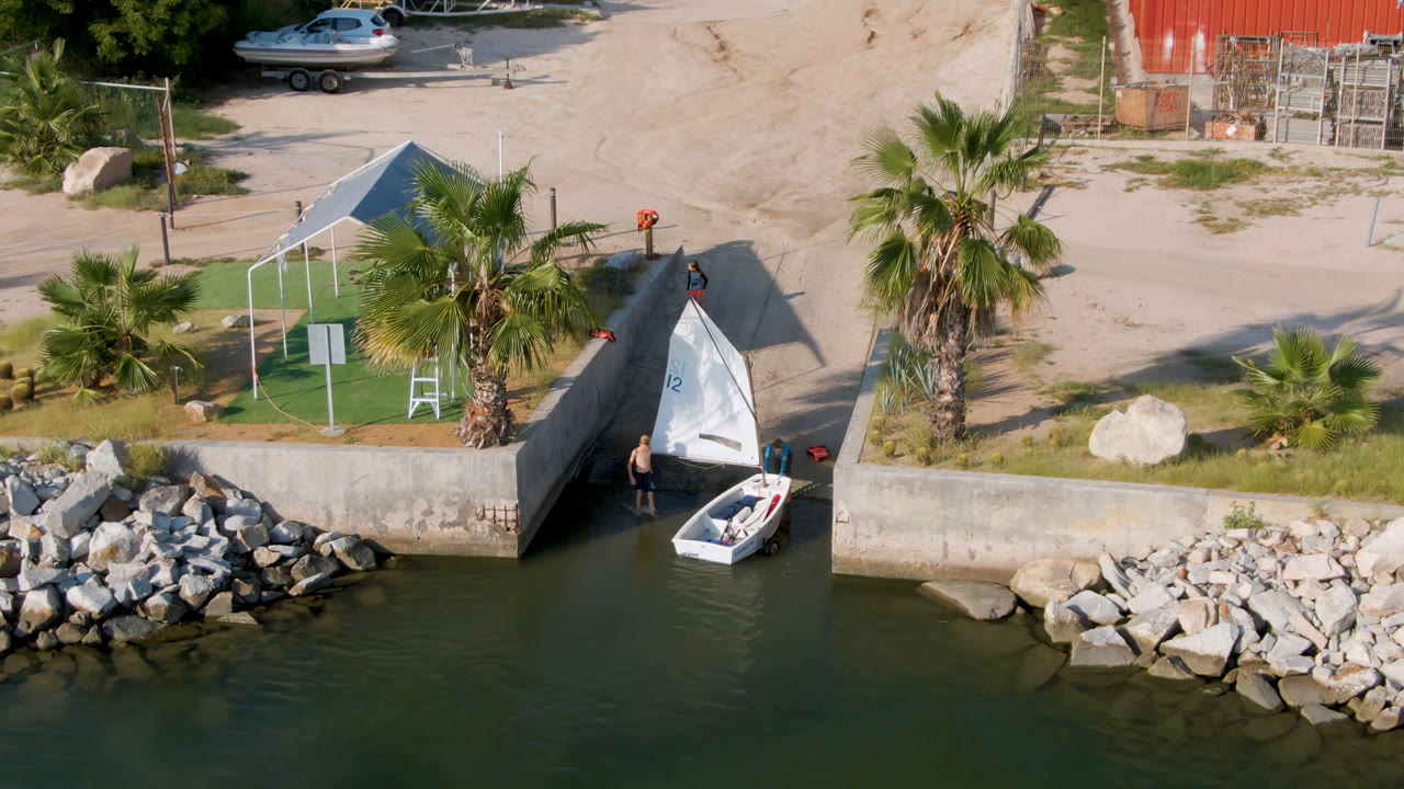 Small sailboat at a concrete ramp beside shoreline rocks and palms.