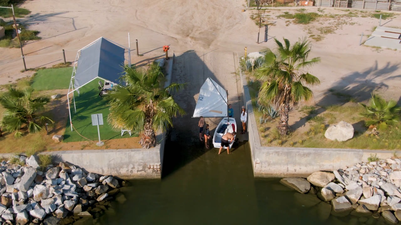 High overhead view in San Jose Del Cabo, Mexico with the sailboat tucked beside the canal wall, the rock breakwater lower in frame, and distant people at the dock.
