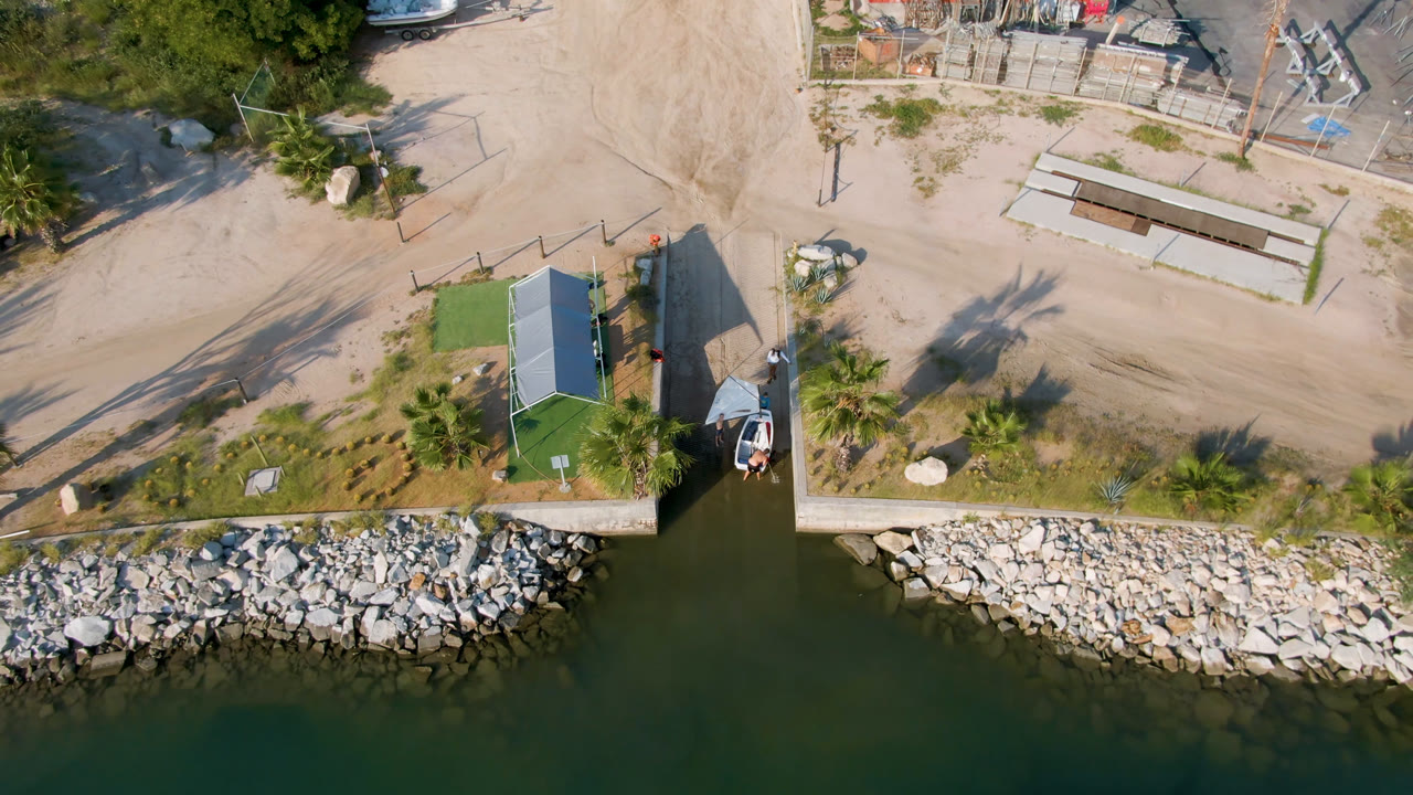 Overhead view in Puerto Los Cabos showing green marina water, a rock-lined shore, a narrow dock, a small boat beside a green-roofed structure, sandy roads, parked cars, and no visible people.