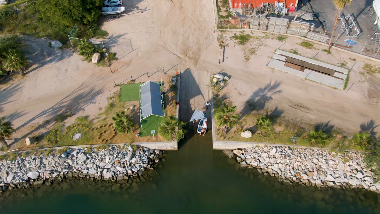Puerto Los Cabos aerial frame with the dock and small boat centered near the shoreline, riprap along the water, sandy access roads, scattered trees, parked vehicles, and no visible people.