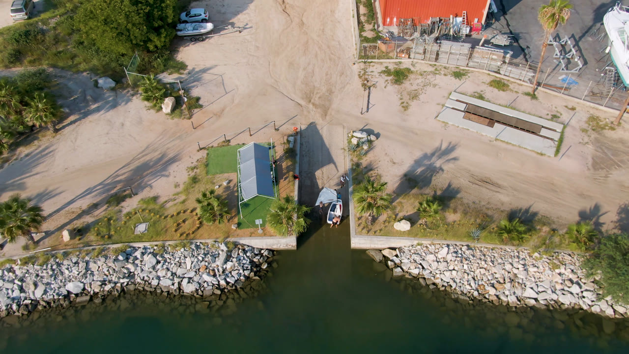 Overhead marina shoreline with dock, small boat, palm shadows, and boatyard area.