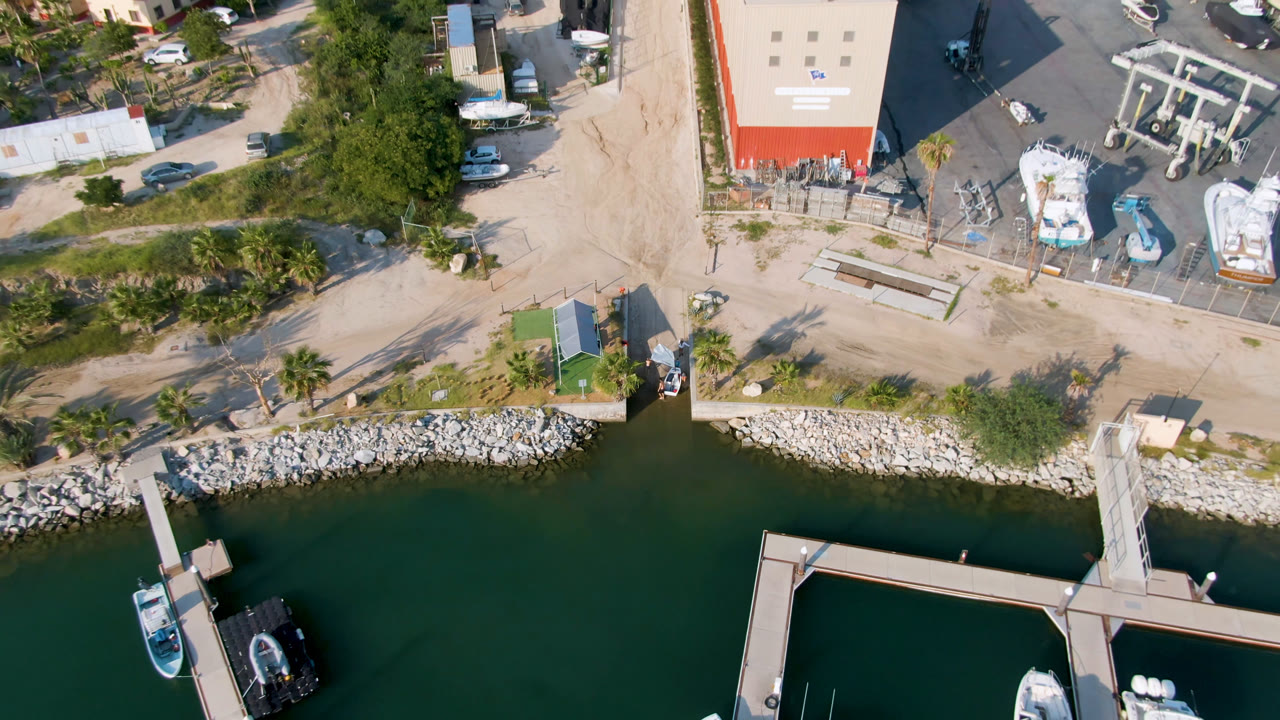 Aerial frame from Puerto Los Cabos showing dark green water below a rocky shore, floating docks with boats, a paved yard, trees, vehicles, and red-roofed buildings, with no visible people.