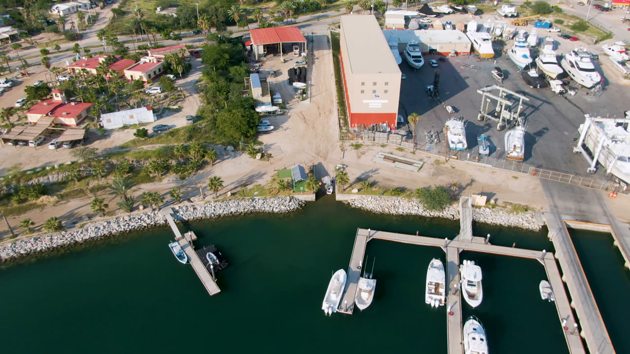 Overhead view of a Puerto Los Cabos marina with dark green water, white boats at floating docks, a rock shoreline, red-roofed buildings, parked vehicles, and no visible people.