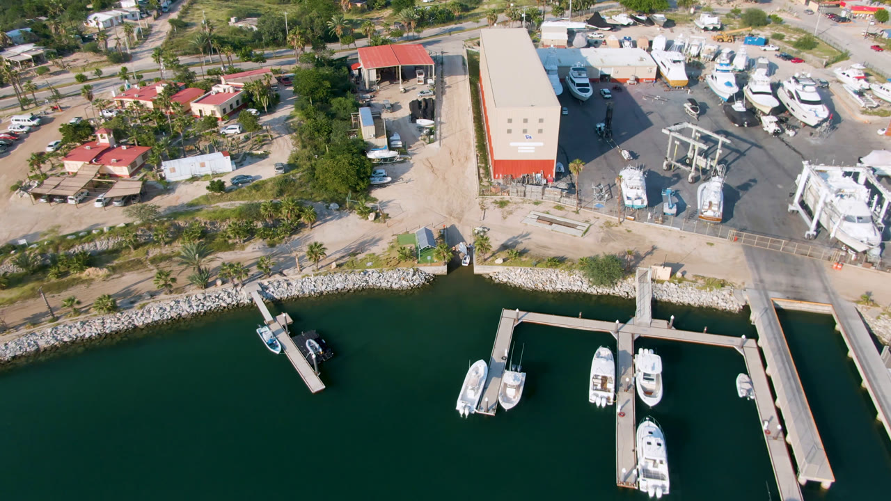 Puerto Los Cabos marina seen from above, with several white boats moored near the lower docks, boatyard equipment and buildings inland, and no visible people.