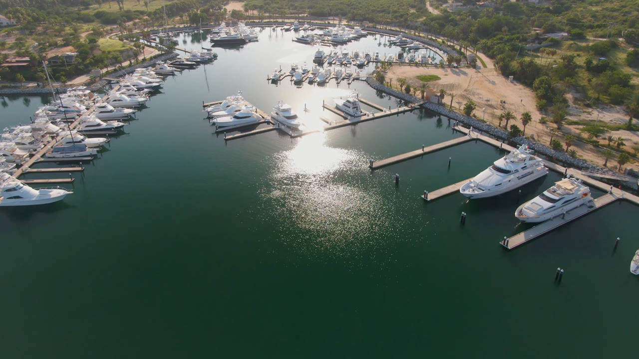 Drone view of Puerto Los Cabos with docked yachts beside piers, a bright patch of reflected sunlight on the water, and no visible people.