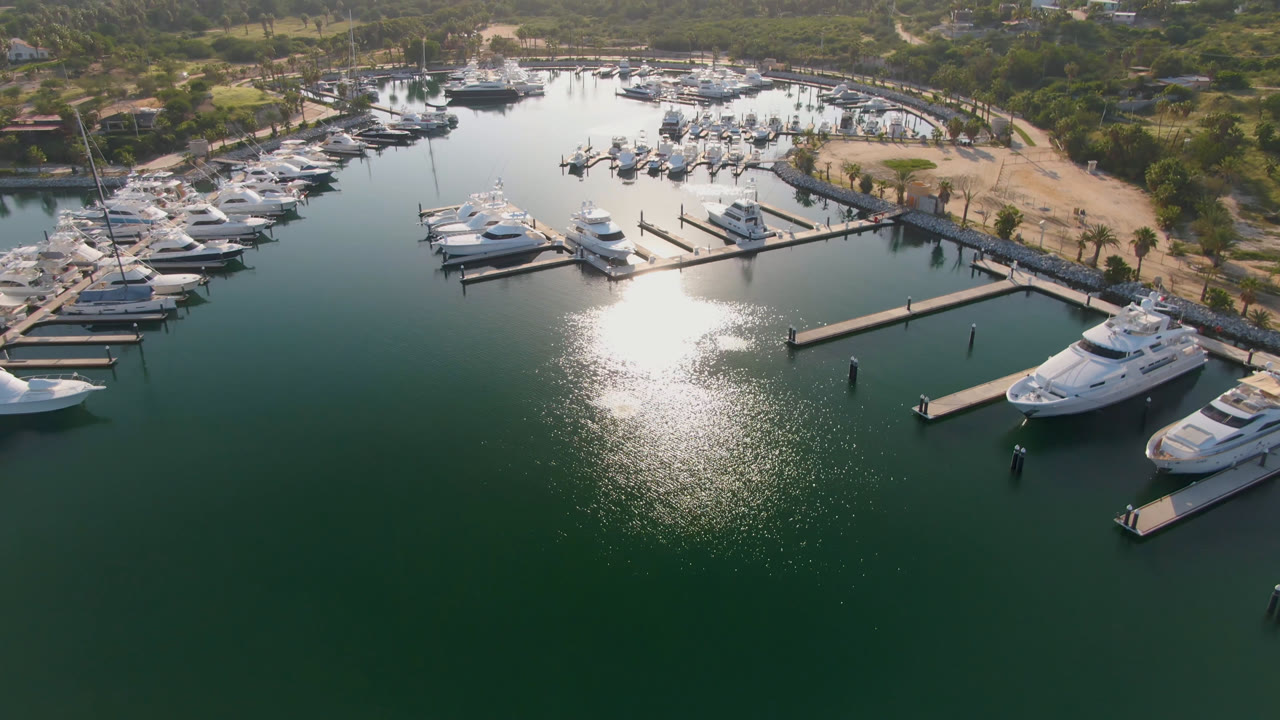 Drone-height view of Puerto Los Cabos marina with the sun glare centered on the water, yachts arranged along several docks, and no visible people.
