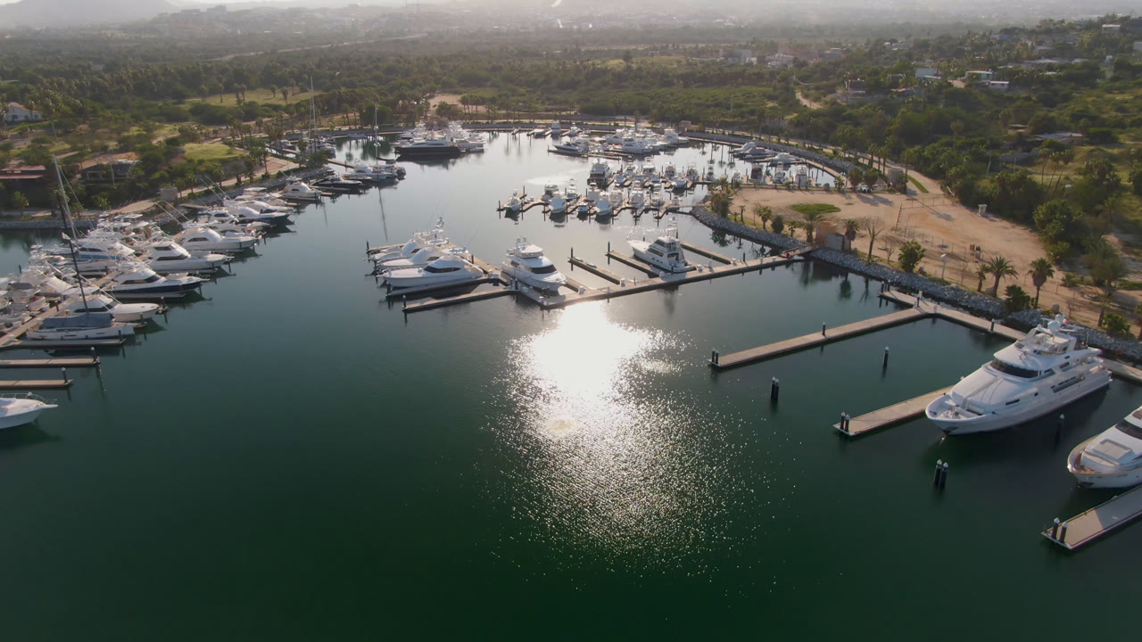 Aerial frame over Puerto Los Cabos marina with white boats along docks, a bright patch of sun on green-blue water, shoreline trees, distant hazy mountains, and no visible people.