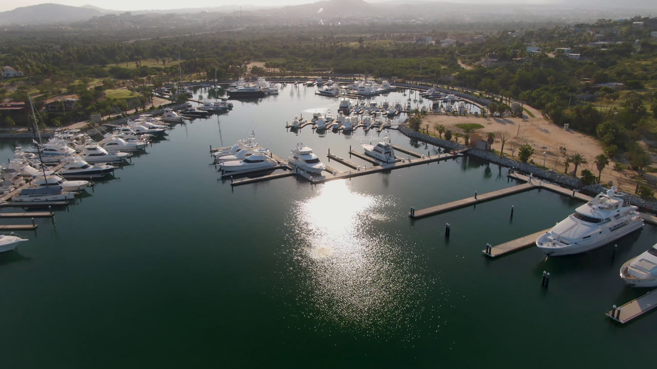 Puerto Los Cabos marina seen from above with yachts clustered at slips, a central dock extending into the water, bright glare on the surface, shoreline roads, palms, and no visible people.