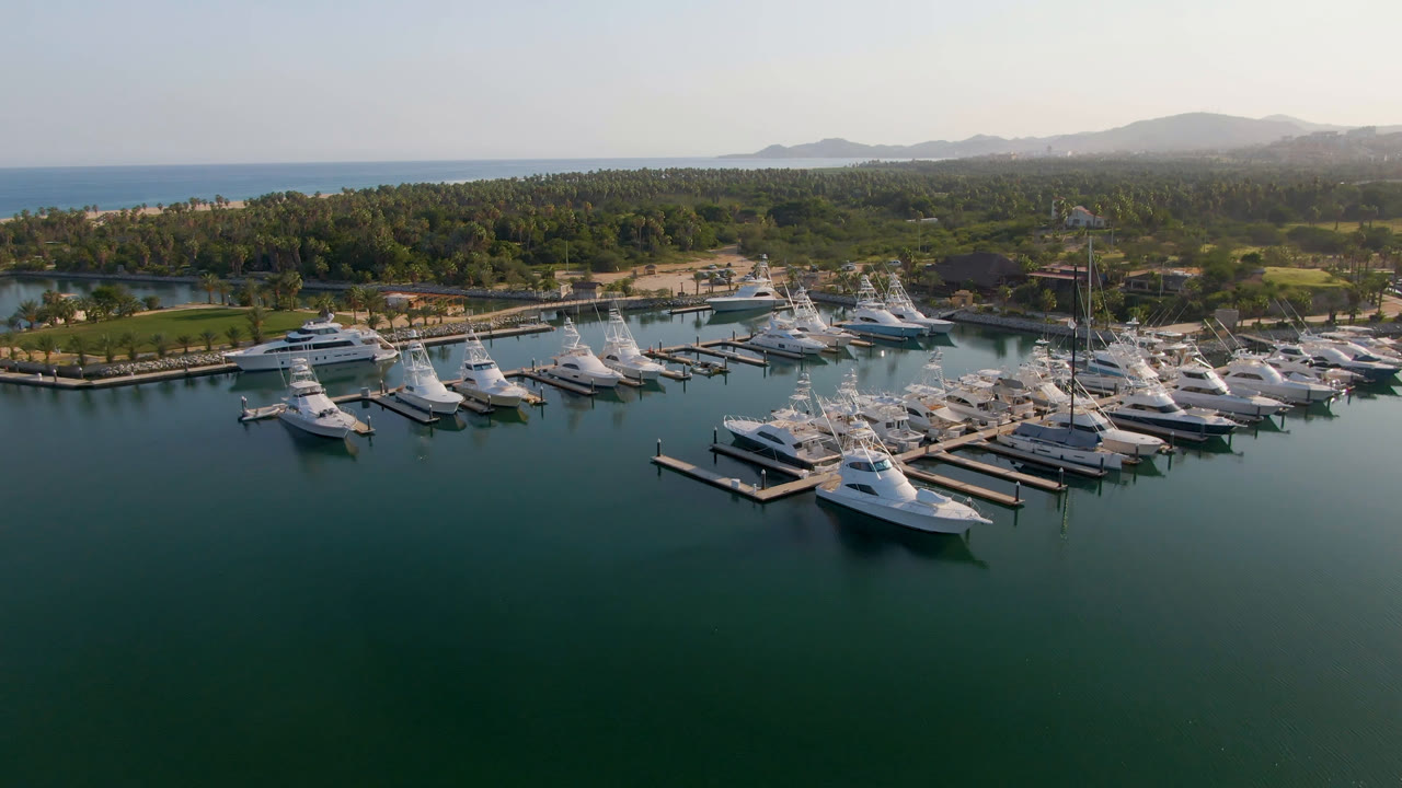 Aerial view over Puerto Los Cabos with marina docks and white yachts centered, open green water below, the ocean band at left, low hills in haze, and no visible people.