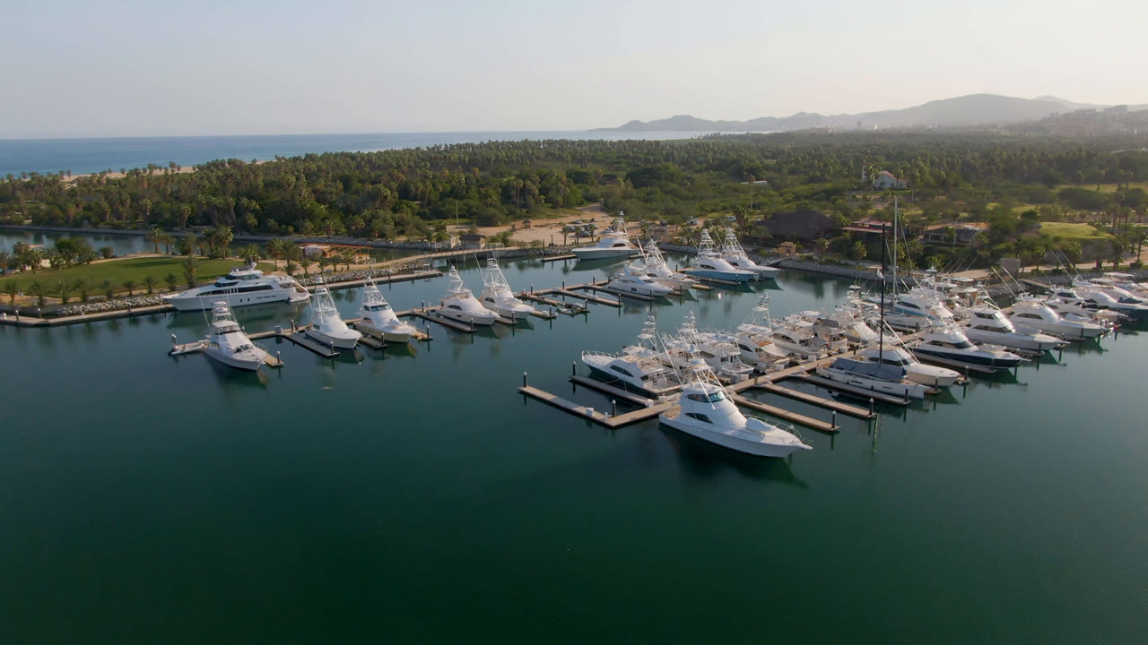 Aerial frame over Puerto Los Cabos marina showing white boats at floating docks, green-blue water in the foreground, palms and hazy hills beyond, and no visible people.