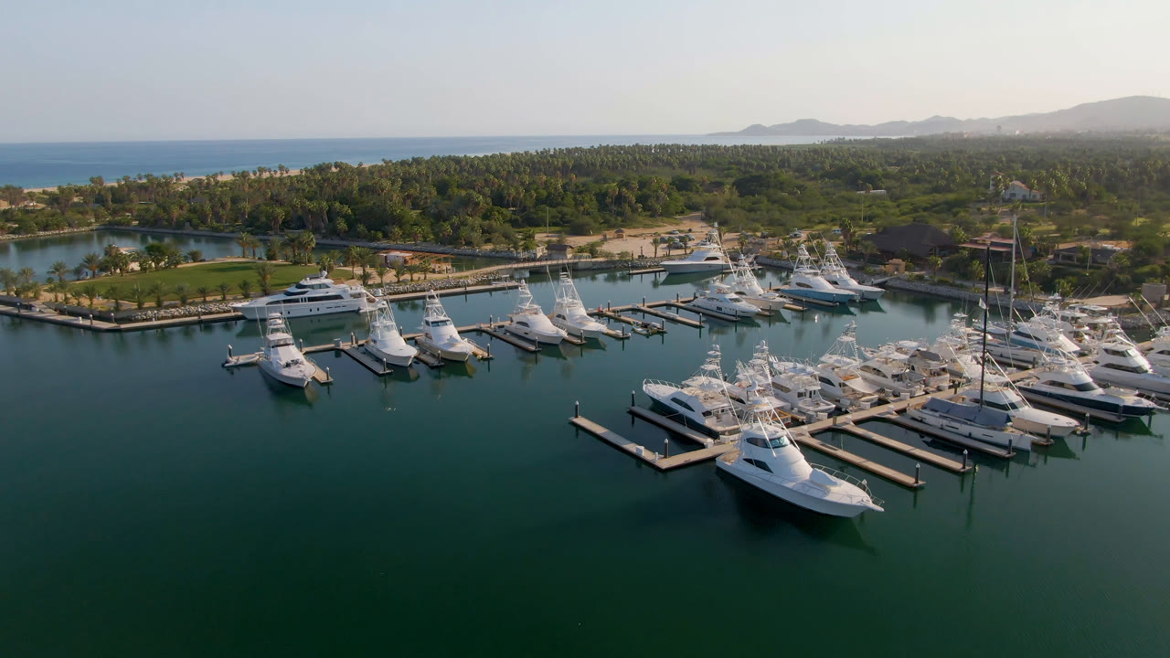 A second aerial frame of Puerto Los Cabos centers the marina basin, with several white boats aligned along docks, a green waterfront area to the left, distant hills, and no visible people.