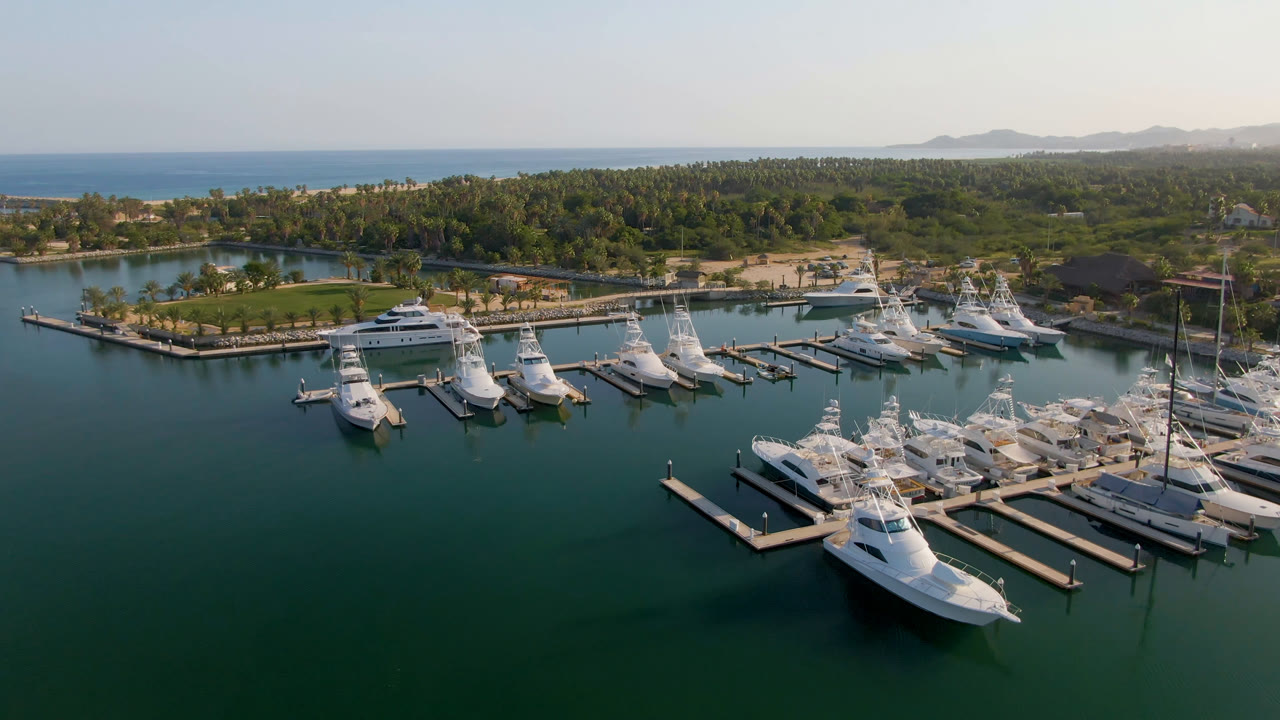 Drone view at 0:01 over Puerto Los Cabos, showing the marina slips slightly farther across the water, with clustered white boats, palms, ocean, and no visible people.