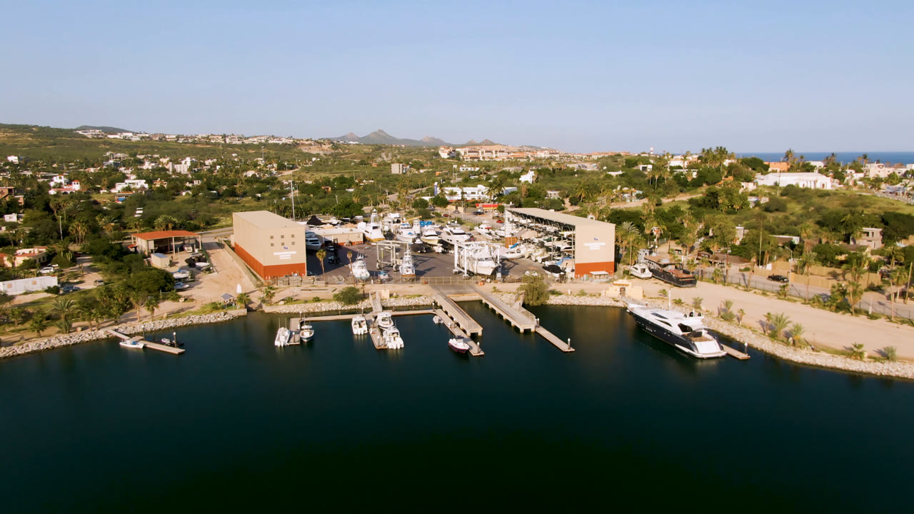 Wide aerial frame of Puerto Los Cabos showing dark marina water, several docks, moored boats, boatyard sheds, parked vehicles, low buildings, distant ocean, and no visible people.