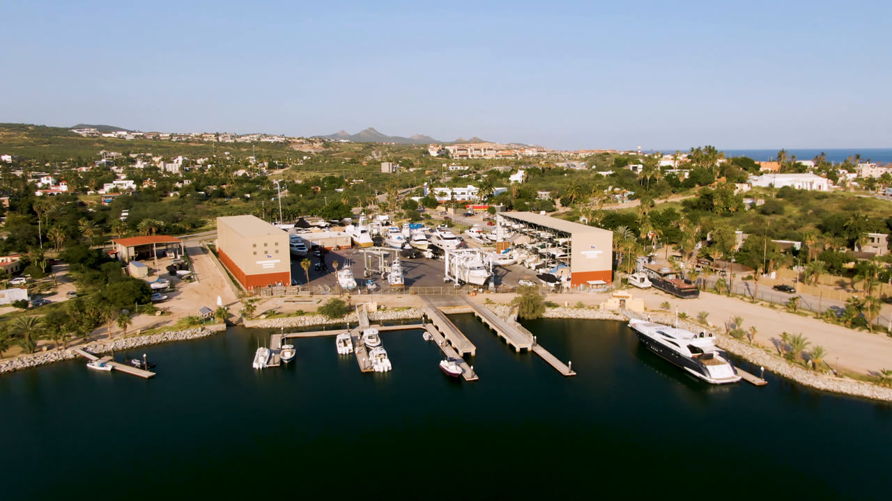 Puerto Los Cabos aerial view with boat slips and a large dock at the water's edge, dry storage areas behind it, hillside buildings beyond, ocean on the horizon, and no visible people.