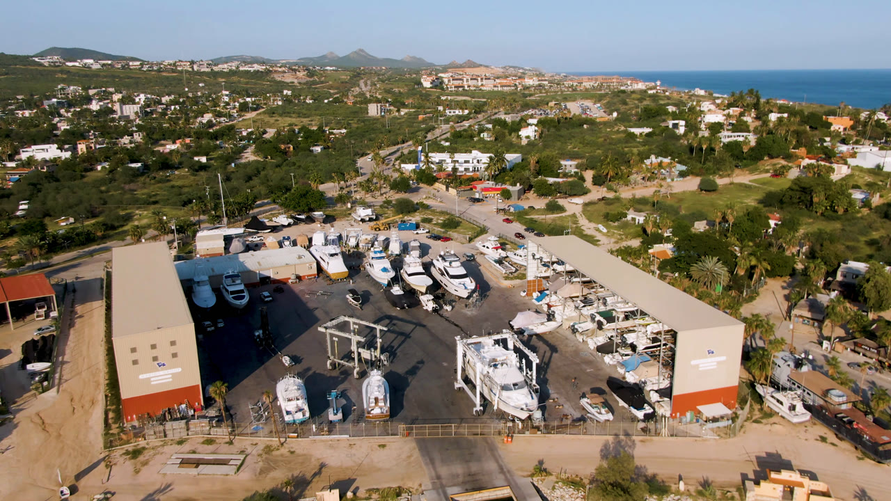 Drone frame in Puerto Los Cabos looking across white boats in a service yard, two long sheds, surrounding roads, green hillside development, ocean, and no visible people.