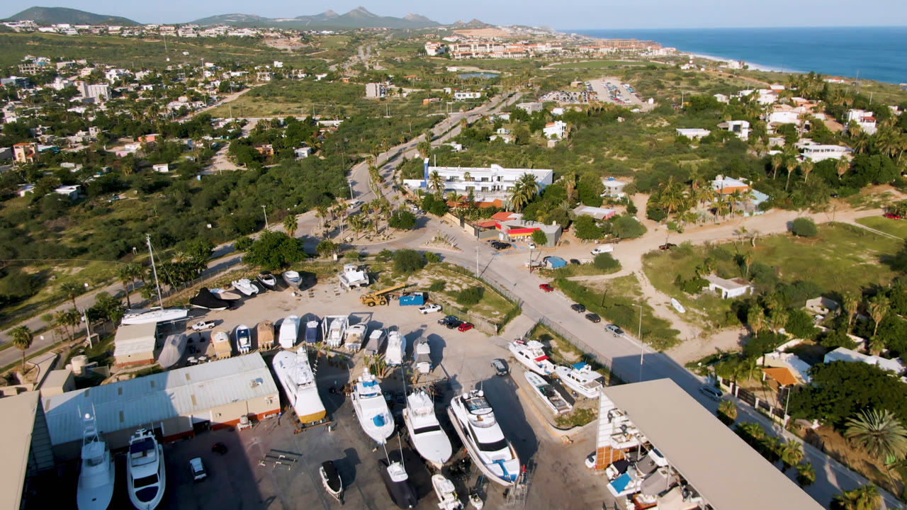 Aerial frame over Puerto Los Cabos showing more of the boat lot centered below, a road cutting through green terrain, distant hills, ocean, and no visible people.