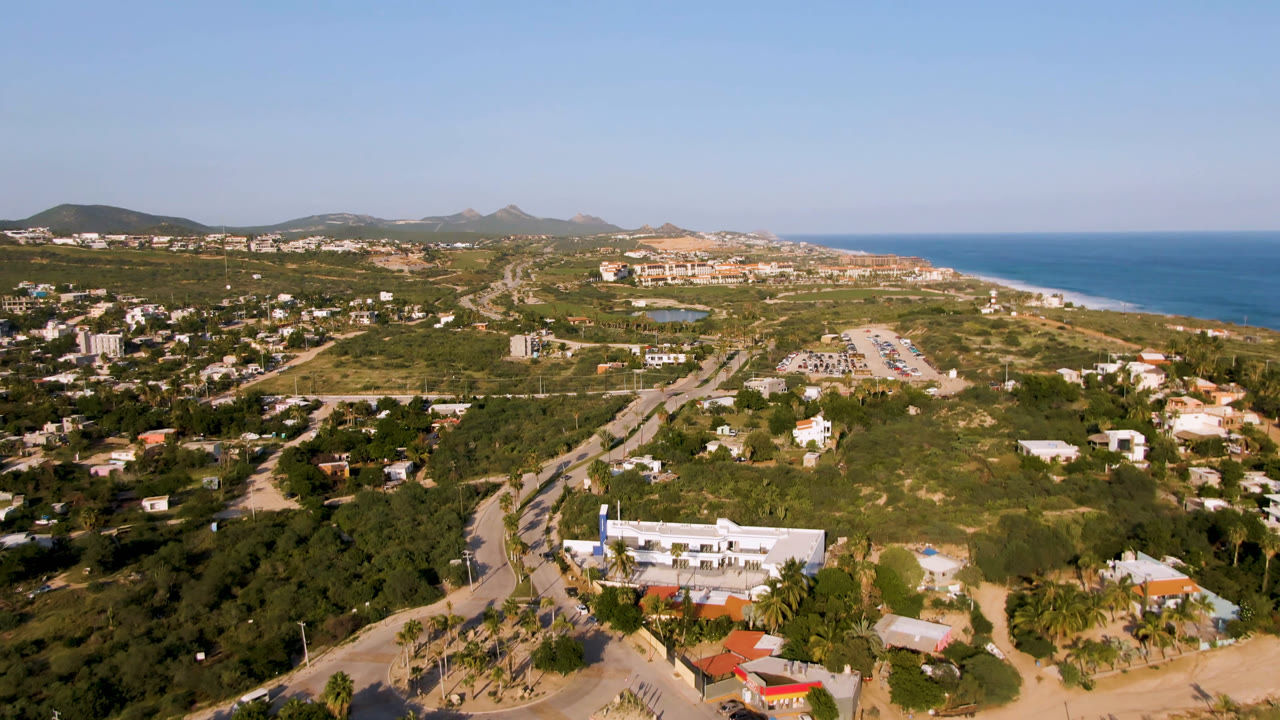 Wide drone view over Puerto Los Cabos, Mexico, with hillside homes, winding roads, palms, sandy beach, blue ocean, distant hills, and no visible people.
