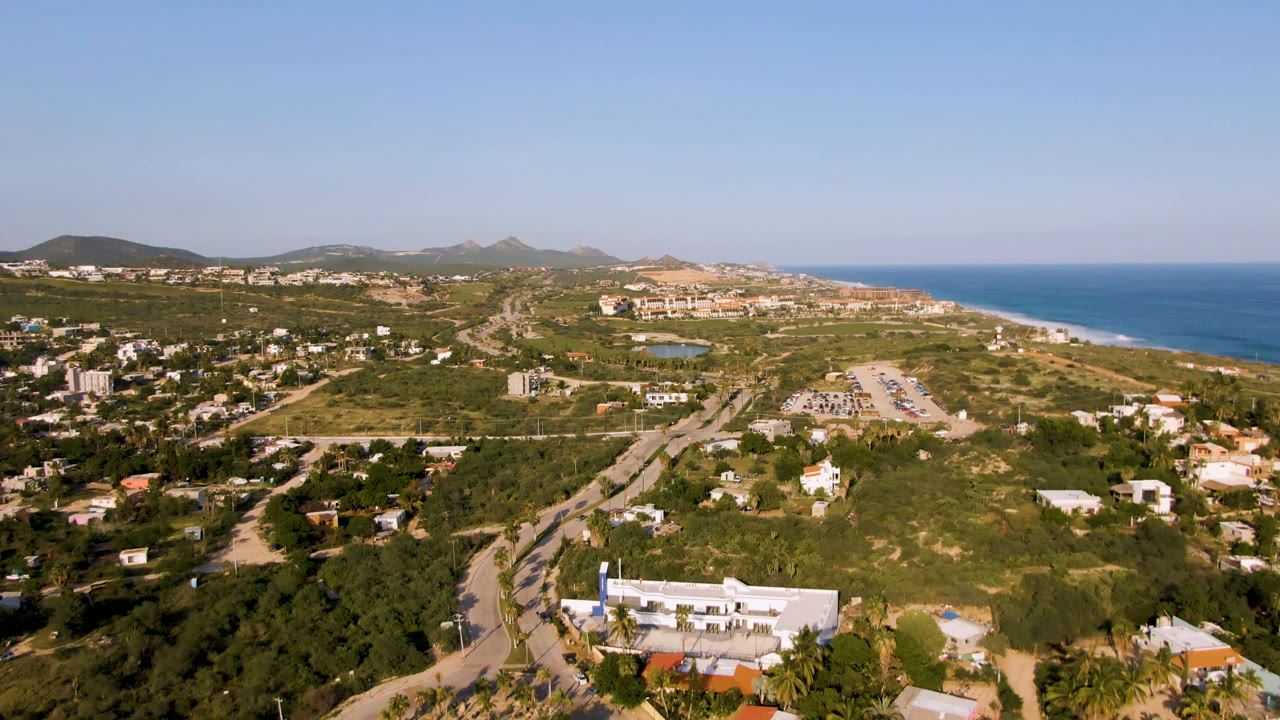 Aerial frame from Puerto Los Cabos showing more of the curving road network, clustered homes, green-brown hills, beach strip, ocean horizon, and no visible people.