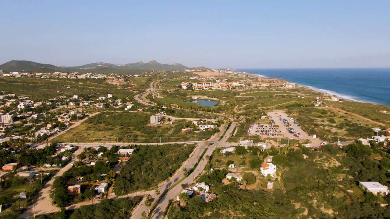 At 0:01, the Puerto Los Cabos aerial holds a broad view of the road network, pale parking area, central lagoon, sandy shore, blue water, low buildings, hills, and no visible people.