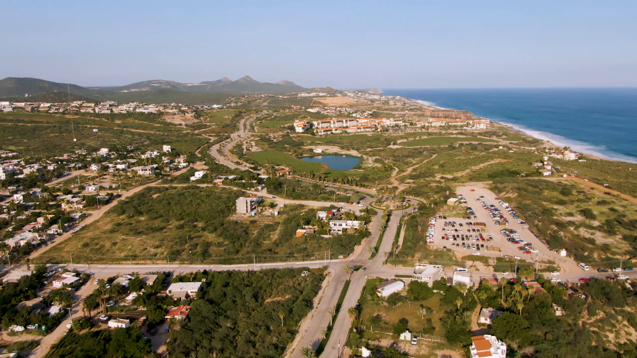 At 0:01, the Puerto Los Cabos aerial frame holds the road grid and parking lot lower in view, with a small blue water feature, beach, ocean, hills, and no visible people.