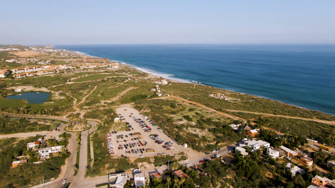 High aerial frame over Puerto Los Cabos with blue ocean at right, surf along the beach, a parking lot and road network below, a small pond at left, low-rise buildings, and no visible people.